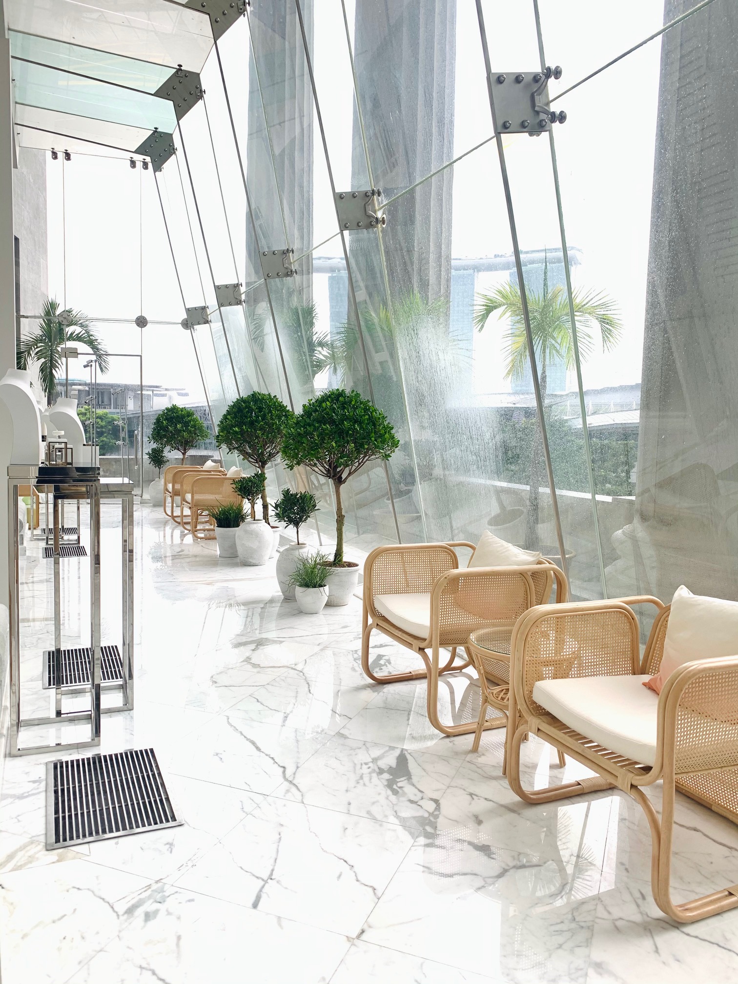 Bright glass corridor lounge with rattan seating, marble flooring, and indoor plants overlooking a modern cityscape