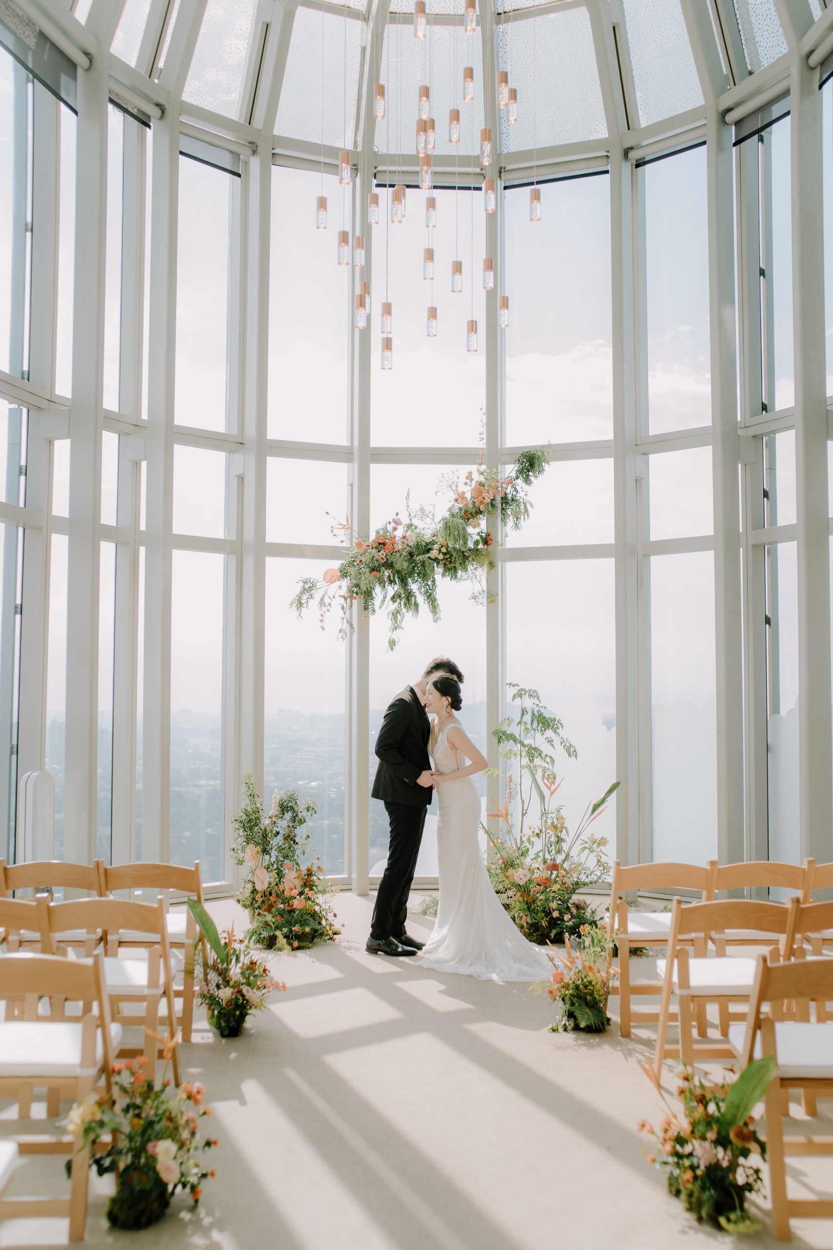 Indoor glasshouse wedding ceremony with couple, floral arch, and modern minimalist seating