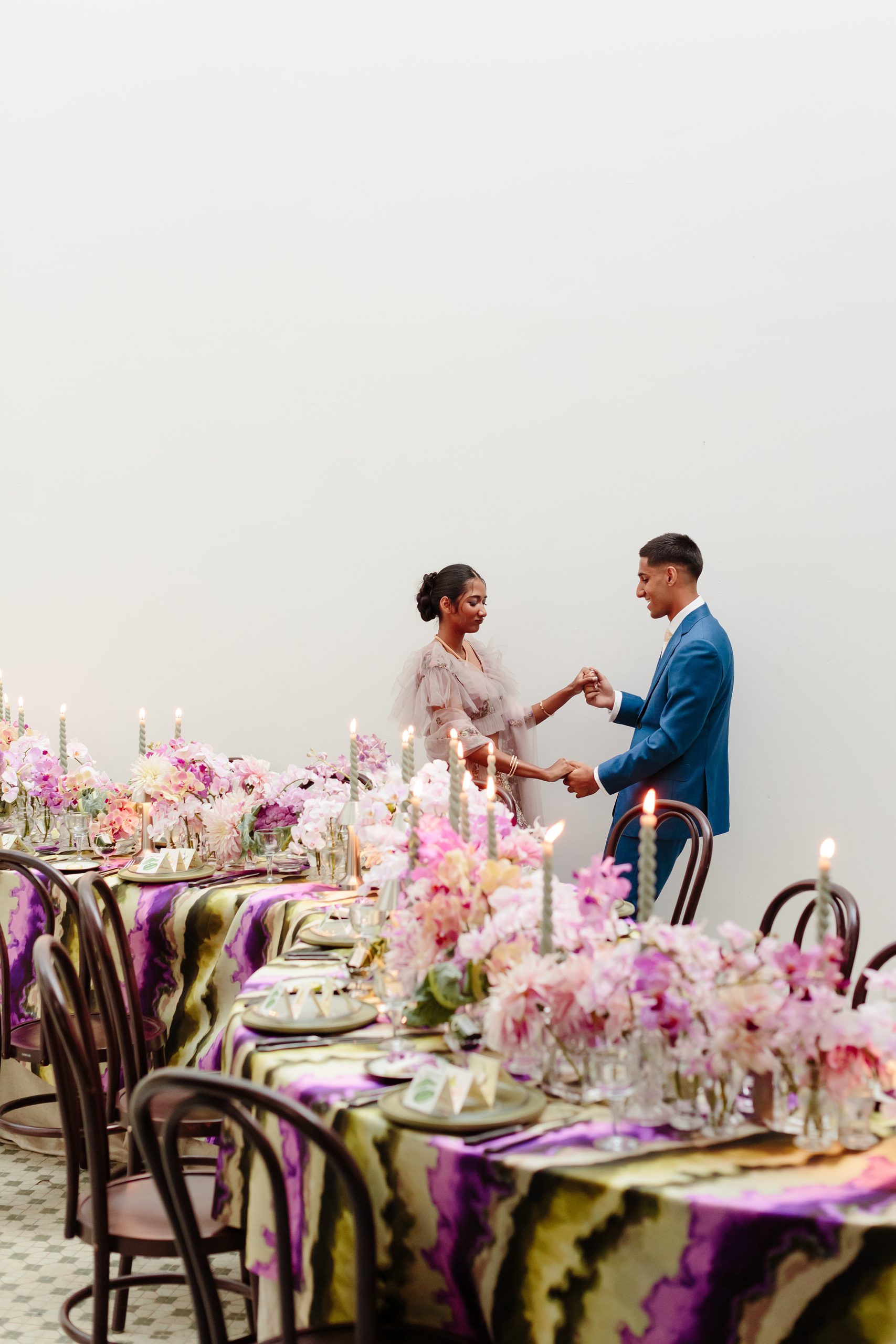 Romantic wedding couple holding hands beside floral table setup with candles and elegant décor